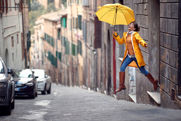 A young girl with a yellow raincoat and umbrella is jumping on the street while walking the city on a rainy day. Walk, rain, city