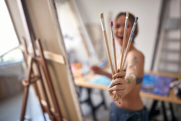 Close-up of a young sexy and attractive female artist in a bra holding bunch of brushes and posing for a photo in her studio. Art, painting, studio