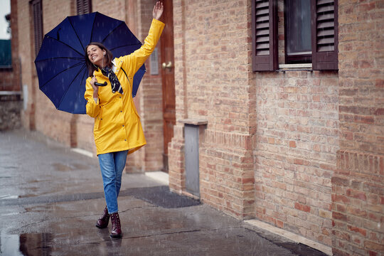 A Young Handsome Woman In A Yellow Raincoat And With Umbrella Is Dancing While Walking The City On A Rainy Day. Walk, Rain, City