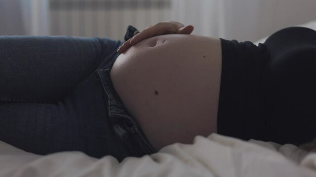 4K footage of a pregnant girl lying on the bed plays with her baby bump and looks how the baby moves inside it
