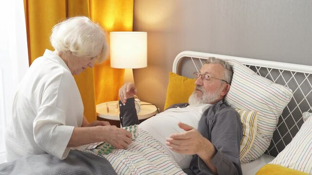 Caucasian Senior man taking medicines and drinking water while lie on bed, wife help and support. Elderly woman take care his husband while he is suffering from flu and high fever temperature