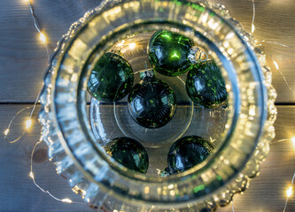 Green christmas balls in a glass on the wooden table with garland lights.
