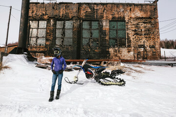 Female rider with snowbike in abandoned winter frozen village. Old fashion russian stile houses in...