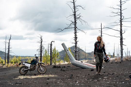 Woman motorcyclist in the dead forest area of active volcano. Post-apocalyptic landscape, The tail of  crashed helicopter, Kamchatka