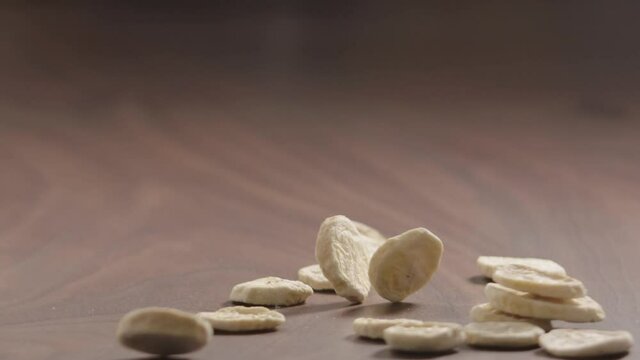 Slow motion freeze dried banana chips fall on walnut wood surface closeup