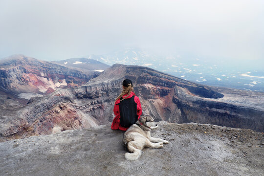 Woman with dog and backpack sitting  on the mountain top above crater and lake of Gorely volcano, Kamchatka