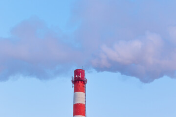 red and white chimney of a factory, from which gray smoke comes out against a blue sky on a sunny frosty winter day