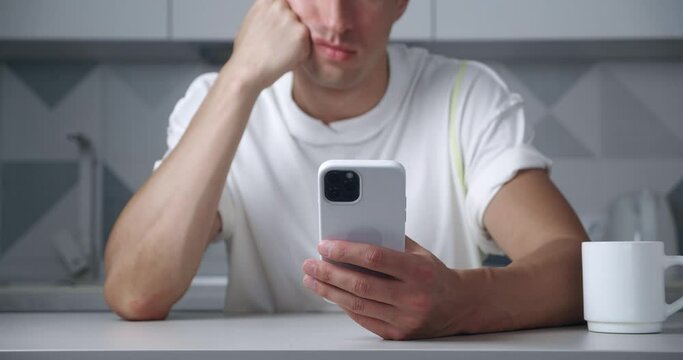 Close-up of bored man surfing internet on smartphone suffering from depression while sitting at home in kitchen. Unhappy male using mobile phone indoors.
