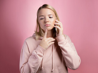 To think about a proposal, a young woman is talking on the phone, thoughtful or doubtful. An attractive young woman in a pink hoodie uses a smartphone, a photo on pink.