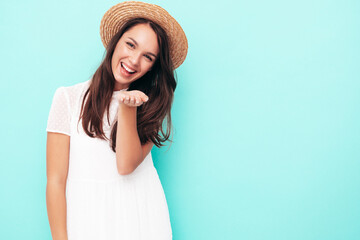 Young beautiful smiling female in trendy summer  clothes. Sexy carefree woman posing near blue wall in studio. Positive brunette model having fun. Cheerful and happy. In hat. Gives air kiss