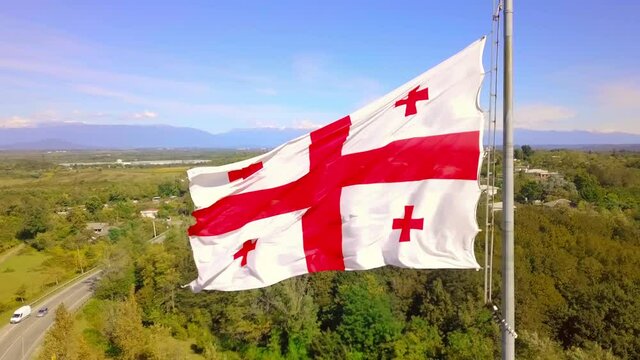 Politics and news of Georgia. Shots with national flag of Georgia against sky from drone. Georgian flag is flying against background of snow-capped mountains and road in village. symbol of Caucasus.