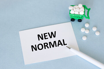 On a light blue background a card with the text NEW NORMAL near the pen and pills in the basket for...
