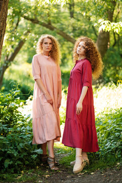 Two young ladies in a summer dresses smiling and posing against in a park. Female portrait outdoors.