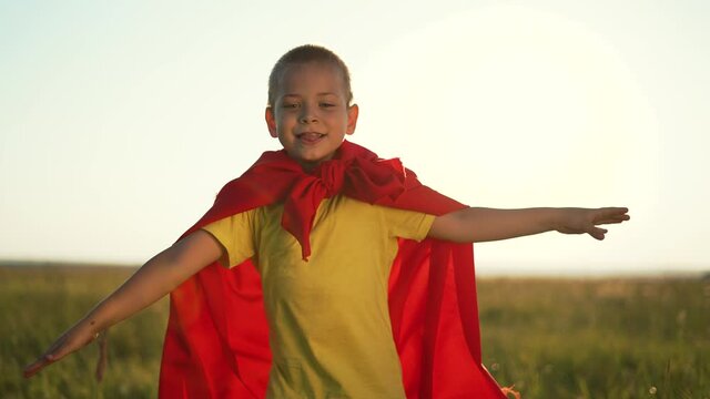 Happy kid in superhero costume. Boy leader run on green grass in park. Childhood dream, child is winner. Boy in red superhero costume in rays of sun. Child hero run on green grass. Child dream in park