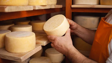Foto op Plexiglas man cheesemaker in the cellar, beautiful wooden shelves with a ready cheese circle, ripening. Cheese production, home basement, indoor. Private entrepreneur. holds in his hands © Valeriia