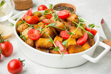 Fried potatoes with fresh cherry and microgreens in a ceramic baking sheet on a light background. Side view, close-up.