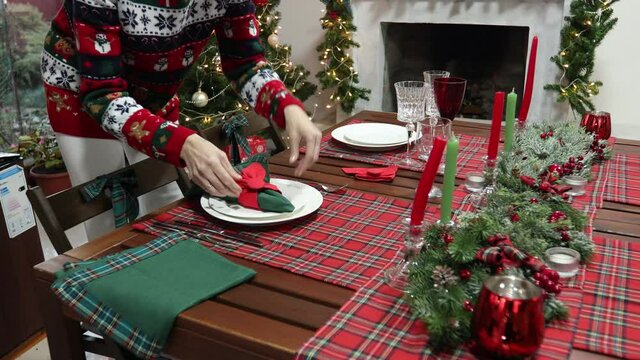 Young woman in sweater with a Christmas pattern decorated the New Year's table against the background of the Christmas tree and fireplace