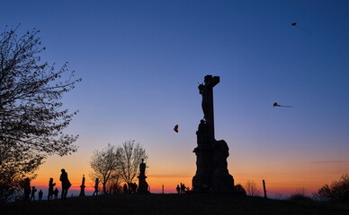 Silhouettes of kids flying kites with stone christianity monument in the foreground at sunset.