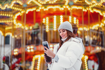 happy girl wears in white down jackets reads smartphone at the Christmas city while holds coffe 