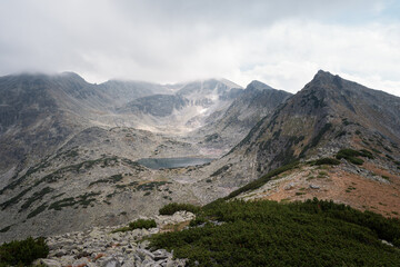 Stunning view towards Musala summit on Rila mountain, over foreground plants, glacier lake and rocky peaks covered in clouds and fog