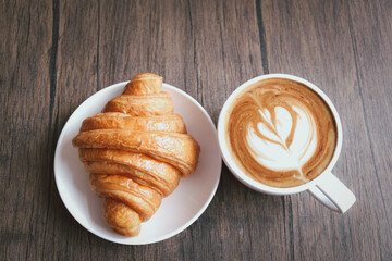 Freshly baked delicious croissant and cup of beautiful morning coffee on wooden breakfast table, top view