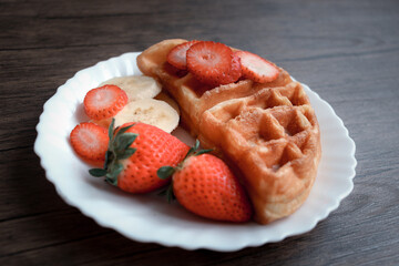 Viennese waffles with strawberry and banana in white plate on dark table.