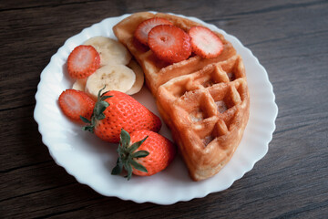 Viennese waffles with strawberry and banana in white plate on dark table.