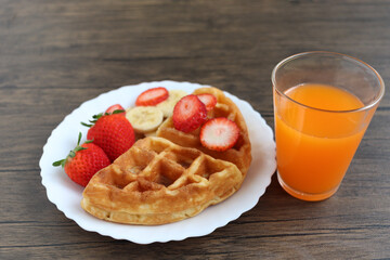 Viennese waffles with strawberry and banana in white plate with orange juice on dark table.