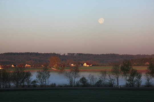 Landscape with fog over Altm&uuml;hl river valley on a spring morning with pale full moon and Hilsberg village in the background, Franken region in Germany