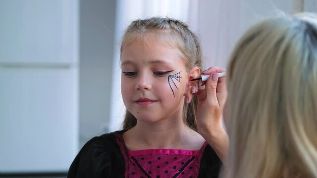 Mom draws a web and a spider on the child's face. Face painting for the holiday of halloween on the face of a little girl dressed as a witch.