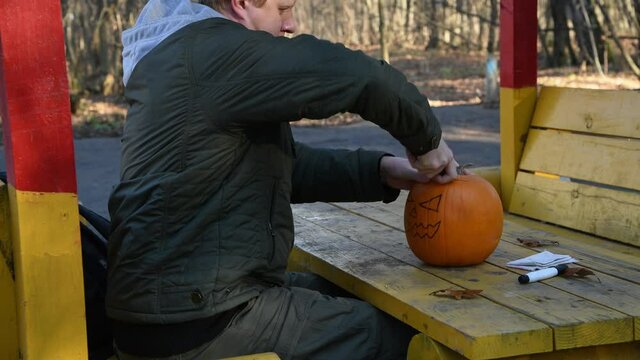 Side view of caucasian man sitting in gazebo outdoor and cutting the top of orange Halloween pumpkin with drawn face on it. Redhead man wears green jacket with grey hood. Holiday decorations theme.