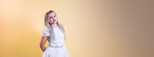 Young and beautiful girl with pigtails on her head and in a white dress stands on a yellow-brown background. Smiles and looks at the camera.
