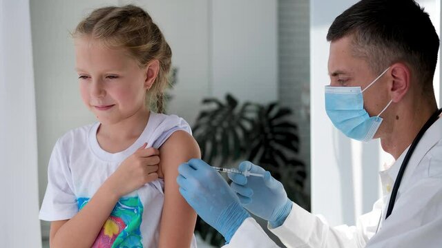 A doctor wearing a medical mask injects a child in the shoulder. Vaccination against coronavirus. COVID-19 vaccine. The doctor vaccinates the kid. A little girl gets a flu shot.