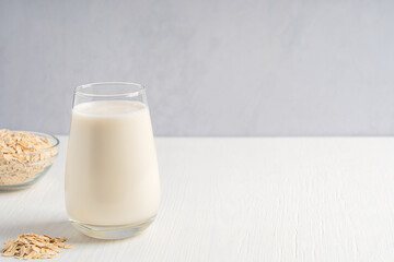Vegetarian homemade plant oat milk with creamy oatmeal taste served in drinking glass on white wooden table for breakfast in the morning with oats grain in bowl. Image with copy space, horizontal