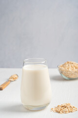 Vegetarian plant based oat milk with oatmeal taste, lactose and dairy free served in drinking glass on white wooden table for breakfast with oats flakes in bowl and spoon. With copy space, vertical
