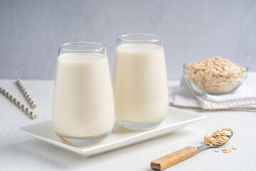 Vegetarian lactose free oat milk made from whole grains with creamy texture and oatmeal-like flavor served in drinking glass on white wooden table with flakes, spoon and paper straws for breakfast