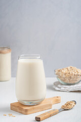 Vegetarian dairy free oat milk made of oats grain with oatmeal flavor served in drinking glass on cutting board with bowl and spoon full of flakes on white table. Image with copy space, vertical