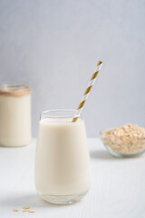 Vegetarian homemade plant-based, lactose free oat milk made of oats grains having sweet oatmeal flavor served in drinking glass with straw and bowl of flakes on white wooden table with jar. Vertical