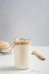 Vegan lactose free oat milk made from whole grains with creamy texture and oatmeal-like flavor served in glass jar on white wooden table with bowl of flakes and spoon. Image with copy space, vertical