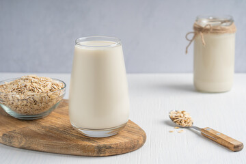 Vegan creamy plant-based, lactose free oat milk made of oats grains having sweet oatmeal flavor served in drinking glass with bowl of flakes on cutting board on white wooden table with spoon and jar