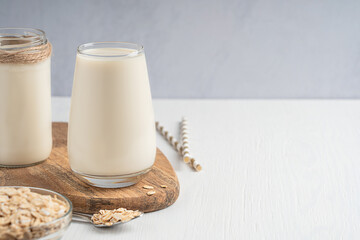 Healthy vegetarian plant oat milk made of oats grain with creamy oatmeal flavor served in drinking glass on wooden board with bowl and spoon full of ingredients on white table. Image with copy space 