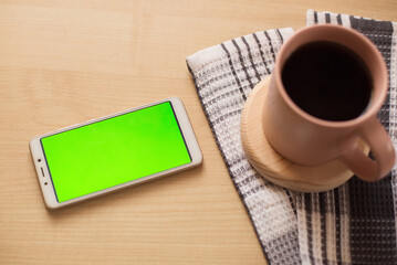 Smartphone with a green screen on the table, next to a cup of tea. Mock up template. Chromakey