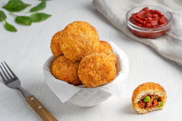 Crispy hot Arancini Italian or Sicilian cuisine rice balls stuffed with minced meat and green peas, coated with bread crumbs and deep fried served in bowl with  fork and sauce on white wooden table
