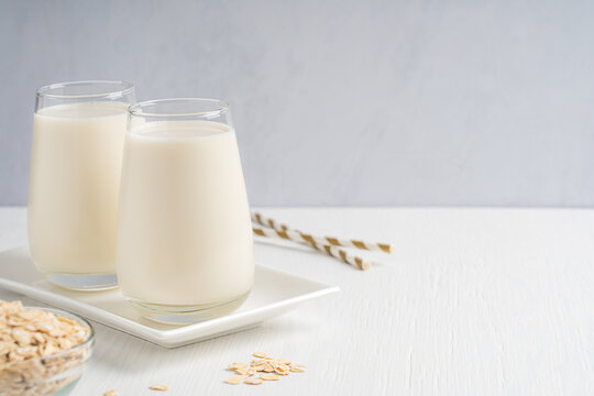 Vegan organic oat milk made from whole grains with creamy texture and oatmeal-like flavor served in drinking glass on plate on white wooden table with bowl of flakes and straws. Image with copy space