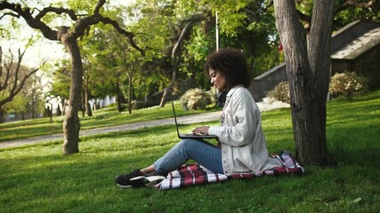 Afro american woman in casual outfit. She smiling, working online on laptop computer while sitting on grass and checkered mat in park