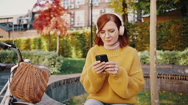Young female hipster sitting on bench in city park, listening to music through headphones and typing on smartphone. Bicycle nearby. Autumn day