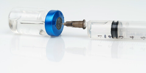 A syringe and a jar of injection lie on a white reflective surface.