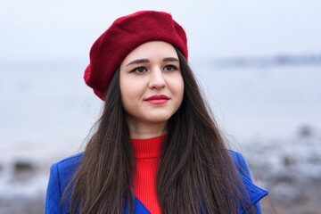 Portrait of beautiful Caucasian girl, young attractive stylish elegant brunette woman with long hair in red beret and gloves and blue coat looking at camera outdoors, walking on beach, ocean. - Powered by Adobe