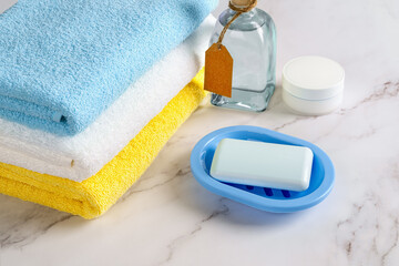 Rectangular soap bar on a blue dish, skin moisturizer and tonic lotion near stack of colored cotton terry towels over marble surface. Washing, hygiene, purity and toiletries concepts.