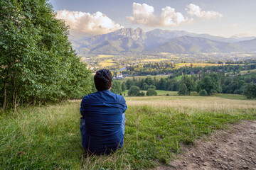 Fototapeta na wymiar View from behind, A male man hiker traveler sitting in the wild looking and admiring mountain and nature. Peaceful and calmness scenic view. Active person outdoor mindfulness living lifestyle concept